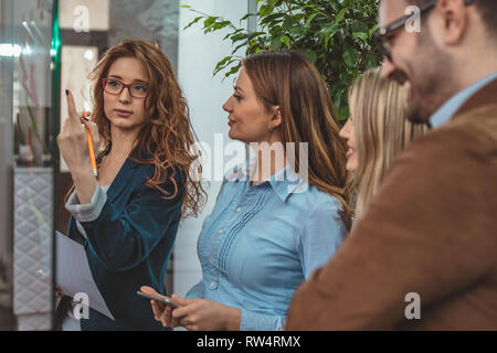 Creative professionals are looking over a post it note wall and discussing and female executive standing at the office behind glass wall with notes Stock Photo