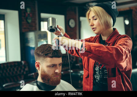 Client during beard shaving in barbershop. Female barber at salon. Gender equality. Woman in the male profession. Stock Photo