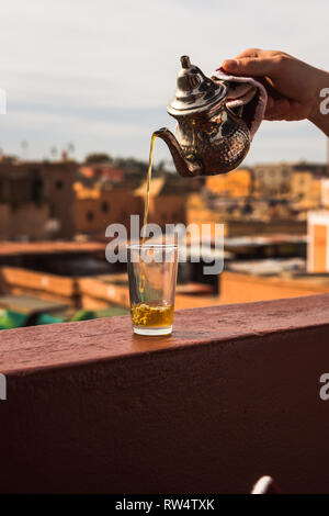 Pouring typical Morrocan fresh mint tea from a silver karraffe into a tea glass above the roofs of Medina of Marrakesh (Marrakesh, Morocco, Africa) Stock Photo