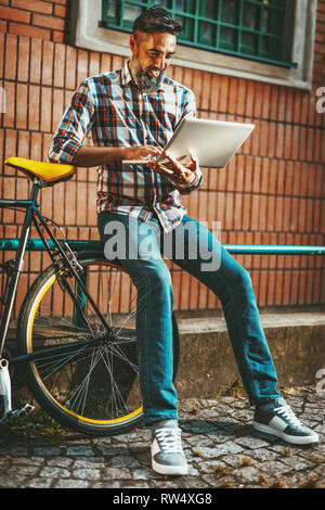 A handsome young man goes to the city with his bike, sitting beside it, waiting for someone and reading some documents on laptop. Stock Photo