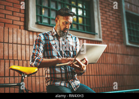 A handsome young man goes to the city with his bike, sitting beside it, waiting for someone and reading some documents on laptop. Stock Photo