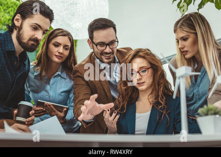 Smart qualified skilled young engineers are looking at the project of alternative energy with miniatures of windmill turbines at the table. Stock Photo