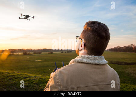 An over the shoulder shot of a male drone pilot hovering and flying his drone in a safe and responsible manner whilst outdoors at sunset Stock Photo