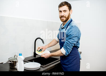 Handsome man in apron doing household chores washing dishes on the kitchen at home Stock Photo