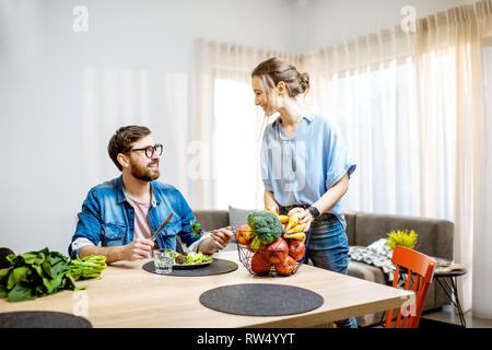 Young man and woman enjoying healthy vegan food sitting at the dinning table full of fruits and vegetables in the modern apartment Stock Photo