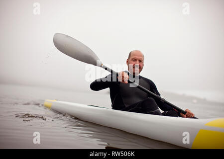 Male kayaker smiles for a portrait as he paddles in the waters of a foggy harbour. Stock Photo