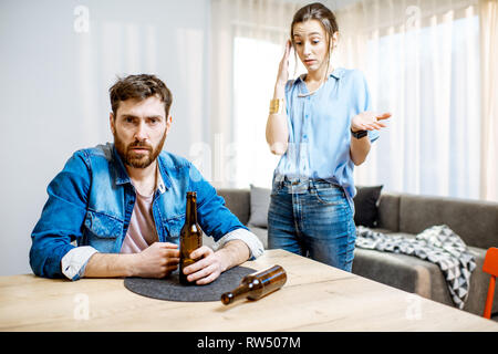Drunk man suffering from alcoholism feeling depressed sitting at home with young woman in despair on the background