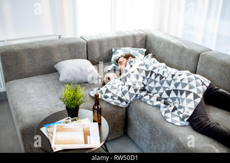 Drunk man lying on the couch covered with blanket, suffering from the alcoholism at home Stock Photo
