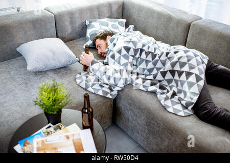 Drunk man lying on the couch covered with blanket, suffering from the alcoholism at home Stock Photo