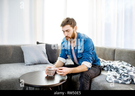 Man drinking some medicines feeling bad or having hangover after the alcohol party, sitting on the couch at home Stock Photo