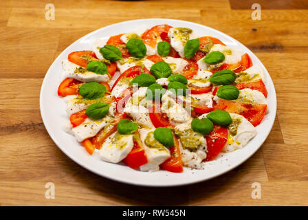 Angled view of plate full of vegan Mozzarella made of cashew nuts with tomato slices, basil and pesto on a wooden table Stock Photo