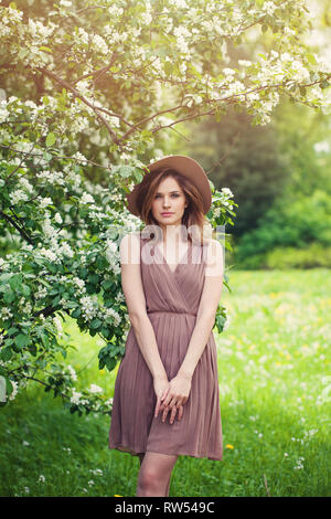 Young perfect woman with brown shoulder-length hair haircut. Beautiful model girl in summer dress and fedora hat on flowers background Stock Photo