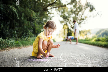 A small cute girl on a road in countryside in sunny summer nature, drawing with chalk. Stock Photo