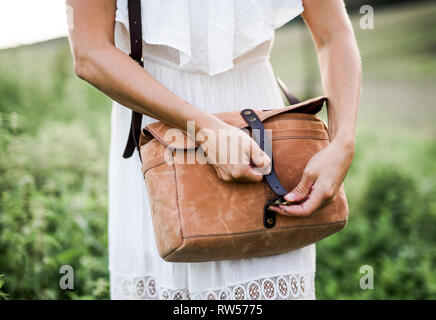 A midsection of woman standing in nature, closing a brown leather bag. Stock Photo