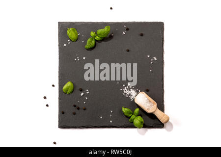 Isolated Slate server on white background with space for text. Arranged salt crystals, peppercorns, wooden scoop and basil leaves. Stock Photo