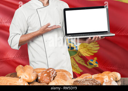 Montenegrin Baker holding laptop on Montenegro flag and breads background. Chef wearing uniform pointing blank screen for copy space. Stock Photo