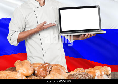 Russian Baker holding laptop on Russia flag and breads background. Chef wearing uniform pointing blank screen for copy space.