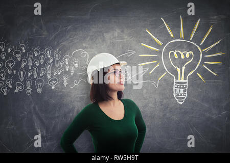 Confident young woman engineer wearing eyeglasses and protection helmet looking away thinking positive. Concept of gathering ideas into a big light bu Stock Photo