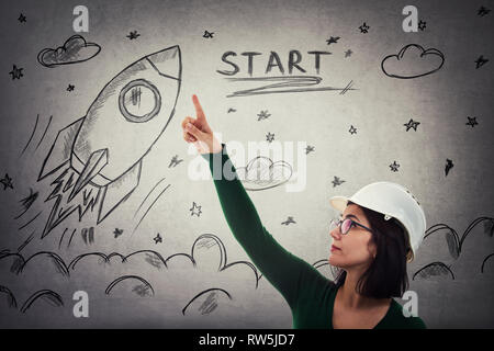 Confident young woman engineer scientist wearing protective helmet and glasses pointing index finger up showing rocket startup sketch, ship taking off Stock Photo