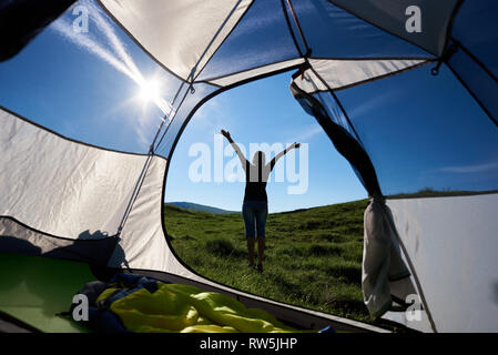 Back view on slim girl hiker lifting hands up in the air, enjoying sunny morning in the mountains. View from tent inside. Lifestyle concept adventure summer vacations outdoor Stock Photo