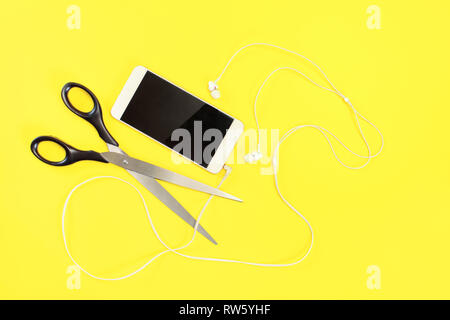 Flat lay photo, white mobile phone with ear buds , scissors about to cut the cable. Yellow background. Stock Photo