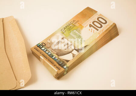 Brown paper envelope next to a stack of ten-thousand dollars in Canadian one-hundred dollar paper currency banknotes on white background Stock Photo