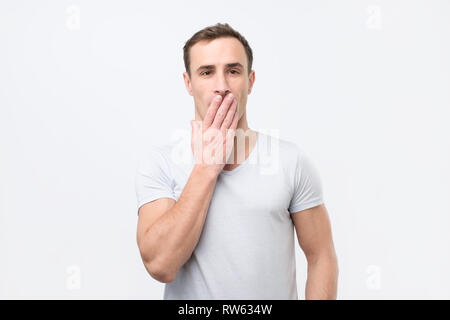 Shocked young italian man in white t-shirt covering mouth with hands and looking at camera while standing against white background Stock Photo