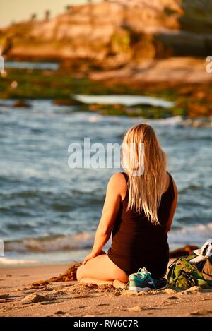 Young, blonde woman sitting and meditating on beach at sunset, at Sunset Cliffs Natural Park, Point Loma, San Diego, California, USA Stock Photo