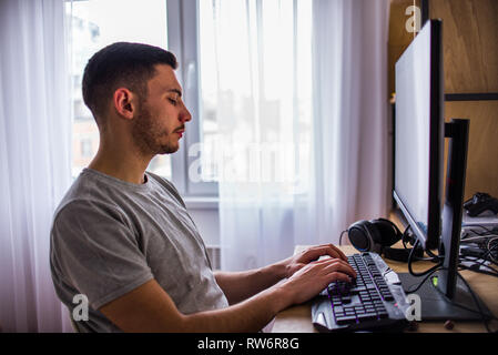 Handsome young man in t-shirt sitting and typing on computer keyboard at home Stock Photo