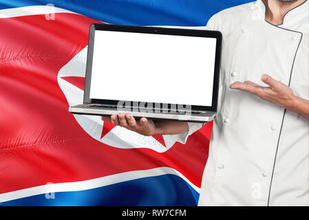 North Korean Chef holding laptop with blank screen on North Korea flag background. Cook wearing uniform and pointing laptop for copy space. Stock Photo
