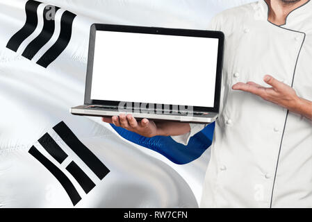 South Korean Chef holding laptop with blank screen on South Korea flag background. Cook wearing uniform and pointing laptop for copy space. Stock Photo