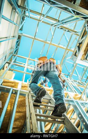 Worker on a Ladder Going on the Upper Level of the Developed House. Skeleton Steel Building Construction Job. Stock Photo