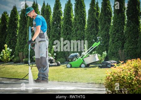 Caucasian Men in his 30s Cleaning Garden Paths and Driveway Using Professional Pressure Washer. Garden Equipment in the Background. Stock Photo