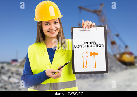 Young female builder smiling as presenting with pen under construction text on clipboard having orange wrench and hammer drawing Stock Photo