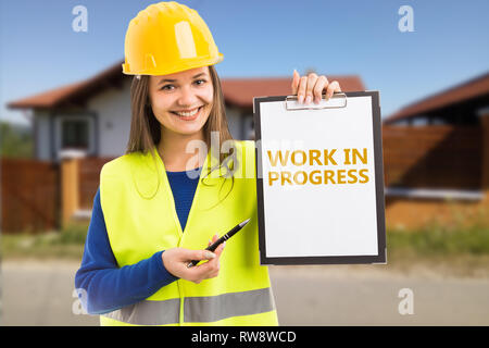Woman builder with friendly expression holding work in progress orange text on clipboard pointing by using pen Stock Photo