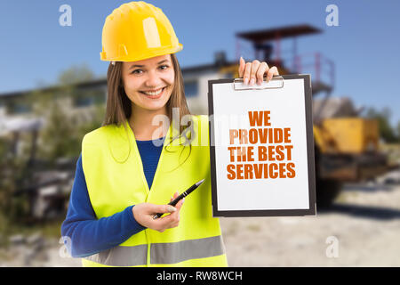 Smiling woman wearing fluorescent vest and yellow safety helmet advertising construction company with we provide the best services text on clipboard Stock Photo