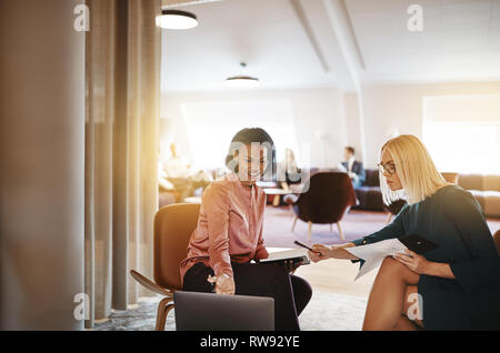Two young businesswomen sitting in a modern office working on a laptop and going over documents together Stock Photo