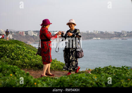 Lima, Peru - February 22 2019: Asian tourists taking photos of the sunset at Malecón de la Costa Verde, woman passing the camera to a friend Stock Photo