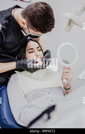 A woman is sitting in a dental office in a dental chair holding a mirror in front of her are dentists. Happy patient and dentist concept. Stock Photo