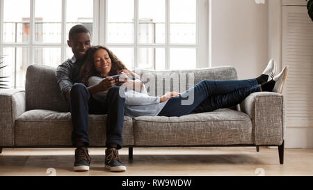 Happy mixed race couple relax on couch using smartphone Stock Photo