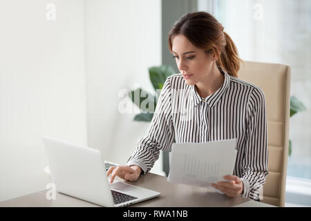Serious focused businesswoman typing on laptop holding papers preparing report Stock Photo