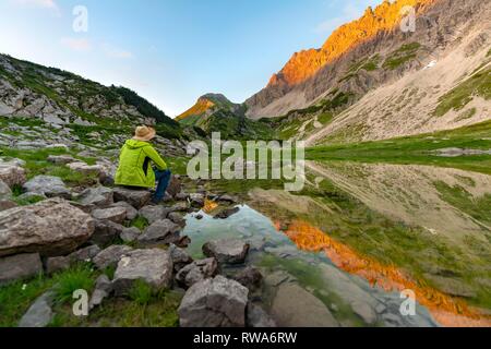 Young man sitting on a stone, reflection in the mountain lake at the Prinz-Luitpold-House, mountains at sunset, Allgäu Alps Stock Photo