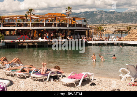 Festival-goers sunbathing and swimming on Zrce beach with mountains in the background during Hideout festival, Croatia, by Georgina Cook Stock Photo