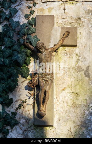 Beautiful rusty Jesus Christ crucifixion statue during sunset partly covered with common ivy, hedera helix on old wall, Olsany cemetery, Prague, Czech Stock Photo
