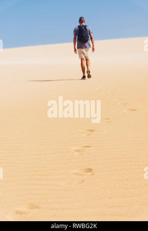 a young caucasian man, seen from behind, wearing a t-shirt and shorts, and carrying a backpack, walking by a white sand desert Stock Photo