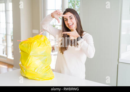 Beautiful young woman taking out the garbage from the rubbish container smiling making frame with hands and fingers with happy face. Creativity and ph Stock Photo