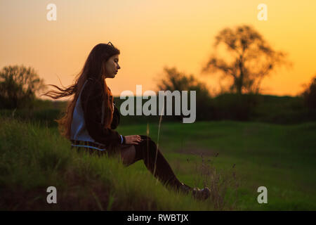 Young girl sitting on grass and contemplate sunset on green spring landscape background in Krivoy Rog, Ukraine. Hair fluttering by wind Stock Photo