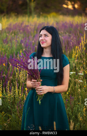 Beautiful happy young girl dressed in marine-colored dress staying at the flowers field and taking bouquet of violet flowers Stock Photo