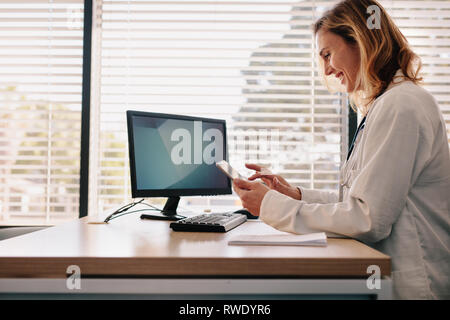 Happy female doctor using her digital tablet in the consultation room. Female physician sitting at her desk and working on her tablet computer in her  Stock Photo