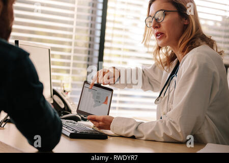 Female doctor talking sharing test results on tablet with her patient. Doctor and patient talking over a medical test result with male patient sitting Stock Photo